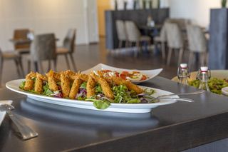 Ein Tisch zeigt zwei Teller mit Essen. Einer hat knusprige frittierte Speisen und einen Salat, der andere einen Salat. Der Tisch ist mit Besteck und Stühlen gedeckt. Der Raum ist hell und geräumig.