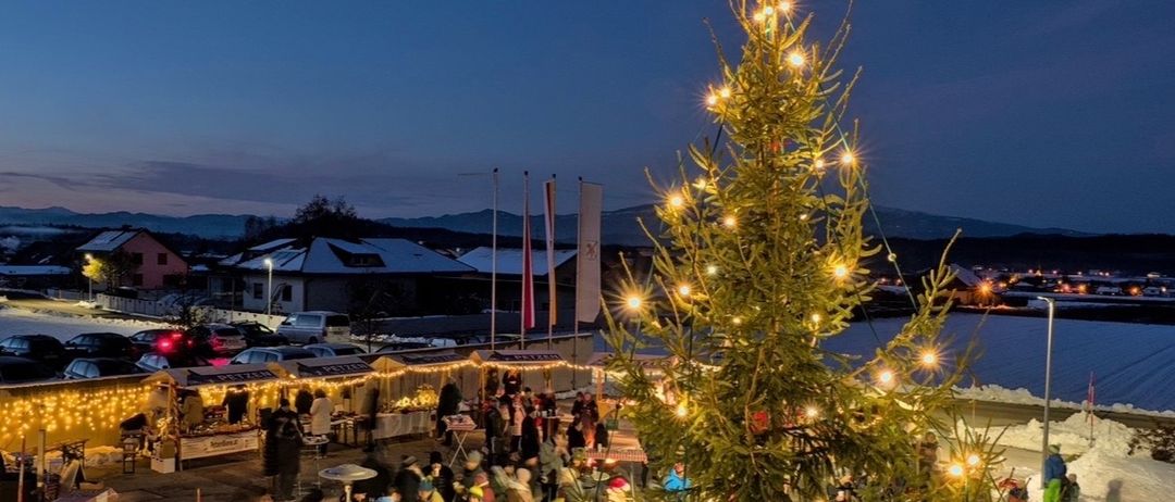 Bei Dämmerung leuchtet ein großer Weihnachtsbaum mit Lichtern inmitten eines geschäftigen Jahrmarktes. Menschen versammeln sich um Stände und Zelte und schaffen eine festliche Atmosphäre.