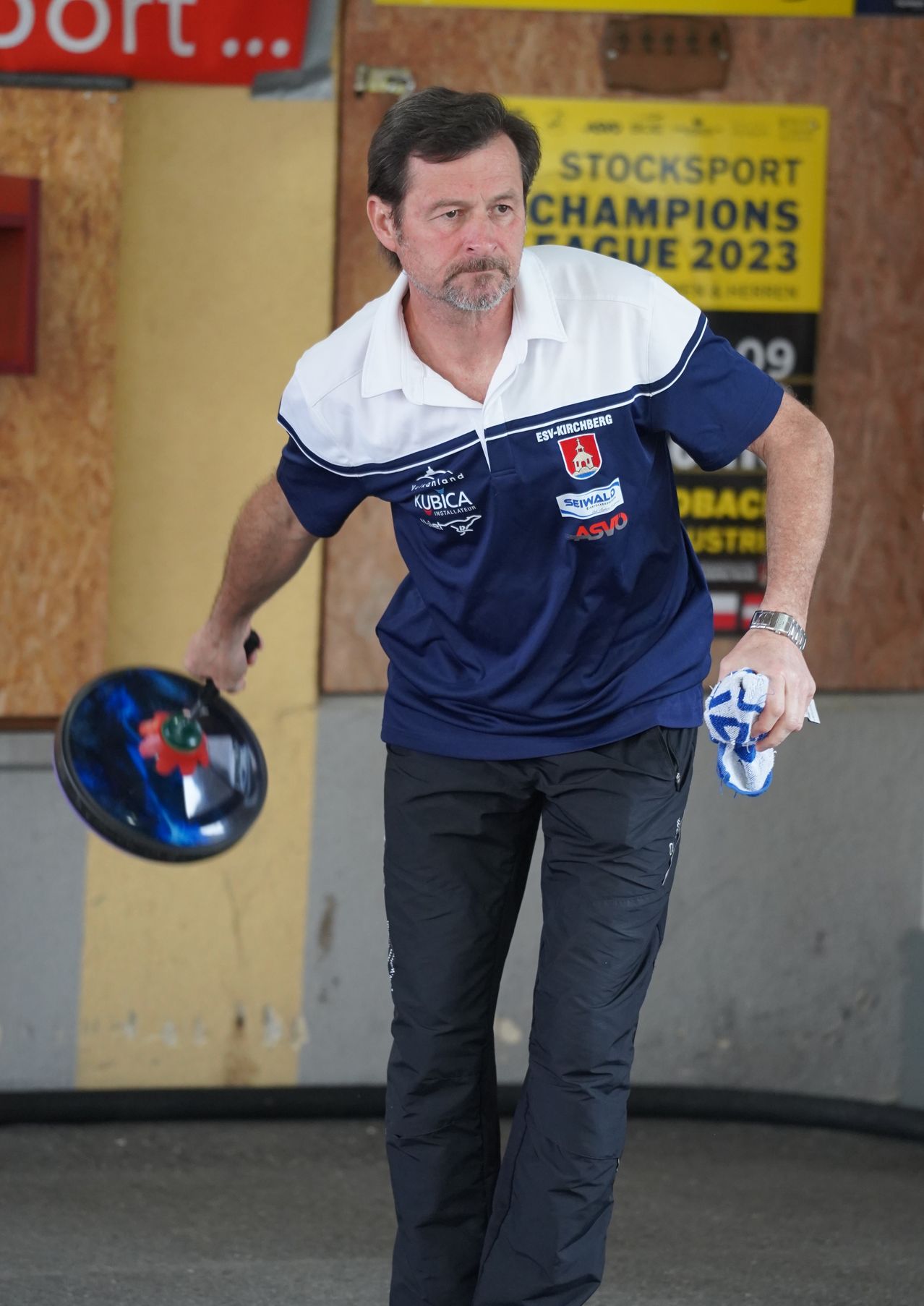 Ein Mann hält einen Bowlingball in der rechten Hand und ein Handtuch in der linken. Er trägt ein blau-weißes Shirt mit Logo und Text. Dahinter hat eine Wand ein gelbes Schild mit den Worten Champions League 2023.