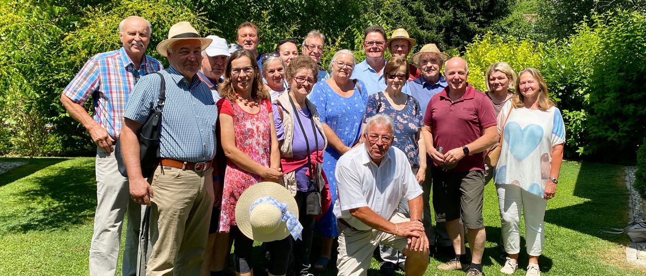 Eine Gruppe von Menschen, darunter Erwachsene und Senioren, posiert für ein Foto im Garten, einige stehend, andere sitzend, mit einem Mix aus Lächeln und ernsten Gesichtern.