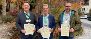 Drei Männer stehen im Freien und halten Urkunden in den Händen, mit Medaillen um den Hals. Sie tragen Jacken und Jeans und lächeln für ein Foto vor einer Kulisse aus Bäumen und Büschen.