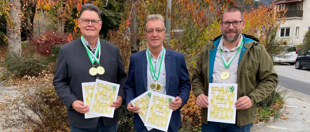 Drei Männer stehen im Freien und halten Urkunden in den Händen, mit Medaillen um den Hals. Sie tragen Jacken und Jeans und lächeln für ein Foto vor einer Kulisse aus Bäumen und Büschen.