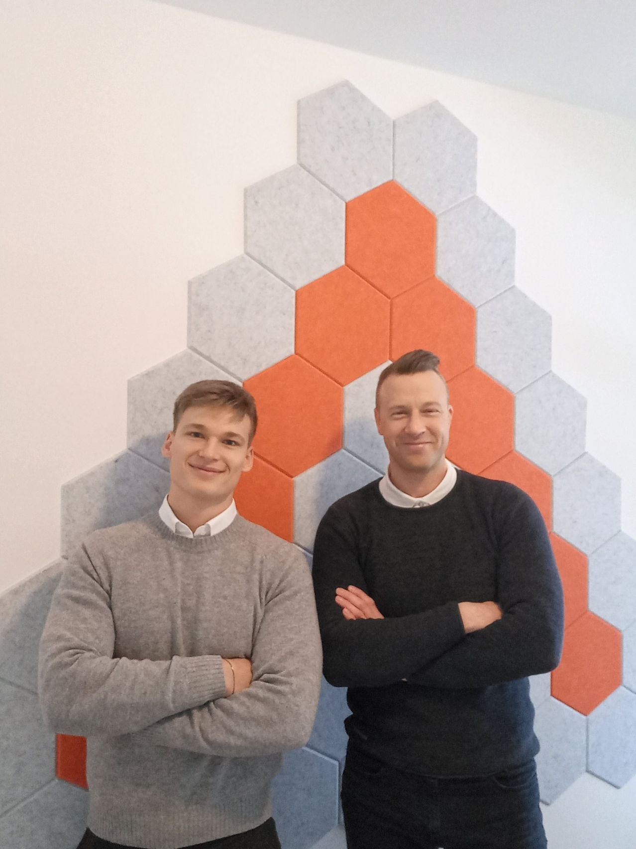 Zwei Männer stehen lächelnd mit verschränkten Armen vor einer geometrischen Wand mit Sechsecken in Orange und Grau.