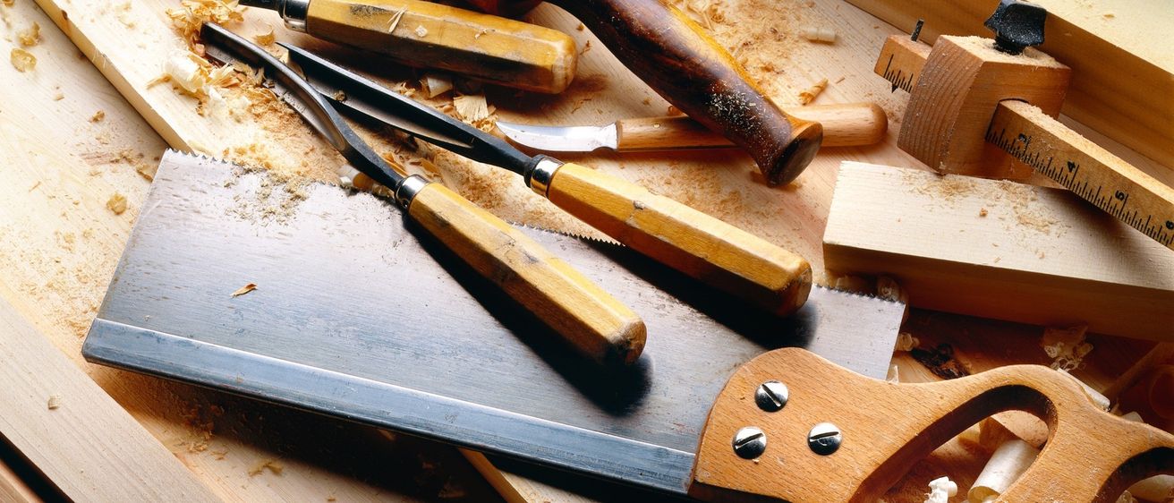 Ein hölzerner Arbeitstisch mit verschiedenen Werkzeugen wie Handsäge, Meißeln und einem Hammer. Sägespäne sind auf der Oberfläche verstreut.