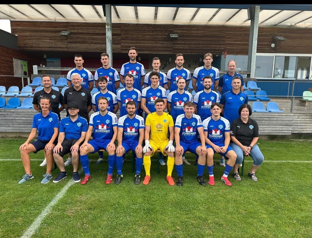Eine Fußballmannschaft posiert für ein Gruppenfoto auf einem Feld mit blauen Stühlen im Hintergrund. Das Team trägt blaue Uniformen mit weißen Akzenten und ein Spieler trägt eine gelbe Uniform.