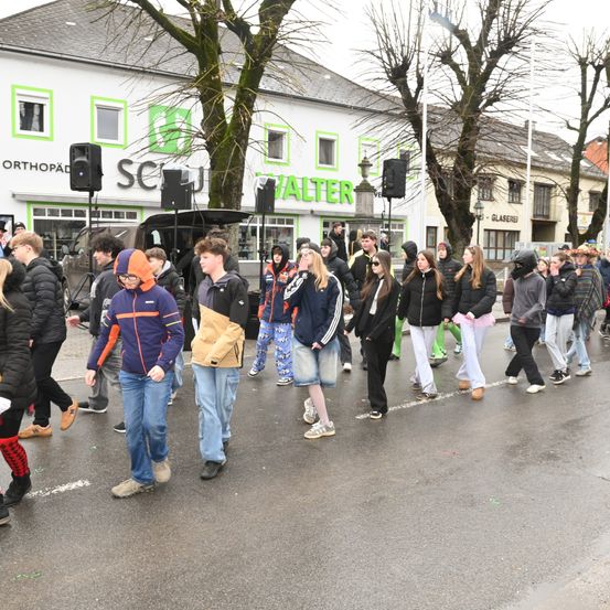 Eine Gruppe von Menschen geht auf der Straße, einige tragen Jacken und Sneaker. Lautsprecher sind vor einem Gebäude mit dem Schild 'SCUL ALTER' aufgestellt. Bäume und ein Auto sind im Hintergrund zu sehen.