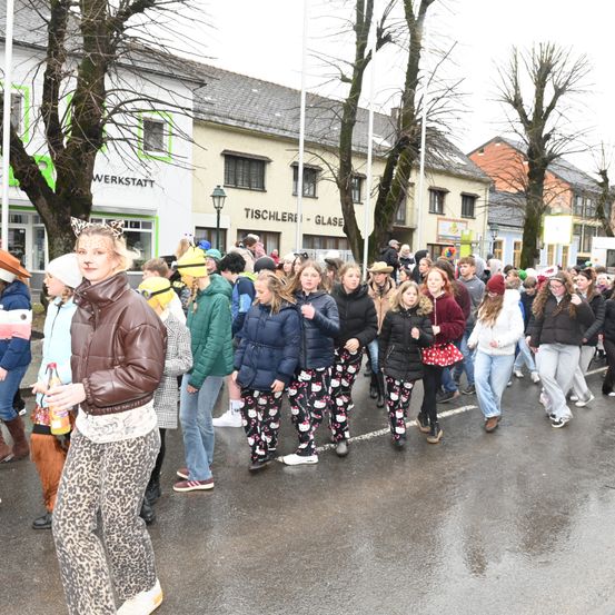 Eine Menschenmenge in Festtagskleidung geht auf einer nassen Straße, vorbei an Gebäuden mit Schildern wie Tischlerei und Werkstatt.