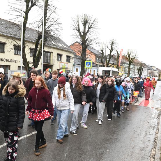 Eine Gruppe von Kindern und jungen Erwachsenen geht in einer Reihe die Straße entlang, in Kostümen gekleidet. Im Hintergrund sind Gebäude mit kahlen Bäumen und ein Fußgängerüberwegschild zu sehen.