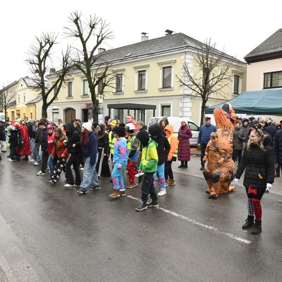 Eine Menschenmenge in verschiedenen Kostümen, einschließlich eines Dinosauriers, steht auf der Straße vor Gebäuden. Bäume und ein grünes Zelt sind sichtbar.