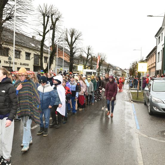 Eine Gruppe von Menschen in Kostümen geht auf einer nassen Straße in einem Umzug. Ein Auto ist am Straßenrand geparkt.