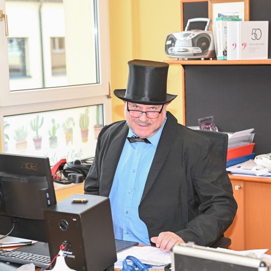 Ein älterer Mann in einem blauen Hemd und einem schwarzen Zylinder sitzt an seinem Schreibtisch und schaut auf den Computerbildschirm. Hinter ihm steht ein Regal mit einem Radio, Büchern und anderen Gegenständen. Ein Fenster zeigt eine Kaktuszeichnung.