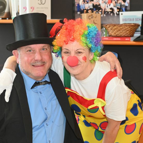 Ein Mann und eine Frau, beide als Clowns verkleidet, posieren für ein Foto. Der Mann trägt einen schwarzen Zylinder und die Frau hat eine farbenfrohe Perücke und eine rote Clownsnase. Sie stehen eng beieinander.