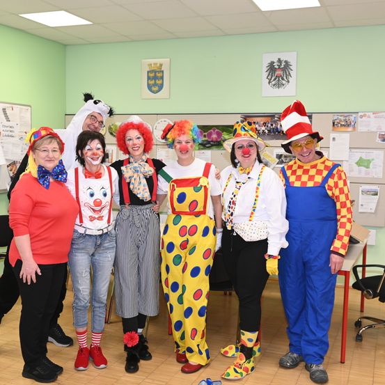 Eine Gruppe von Menschen, die als Clowns verkleidet sind, posiert für ein Foto in einem Raum mit grünen Wänden und verschiedenen Plakaten. Sie tragen bunte Kostüme und Schminke.