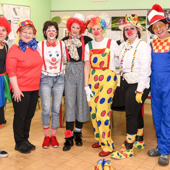 Eine Gruppe von Menschen, die als Clowns verkleidet sind, in einem Raum, einschließlich einer Person in einem roten Clownskostüm und einer anderen in einem blauen Clownskostüm. Sie alle lächeln und posieren für ein Foto.