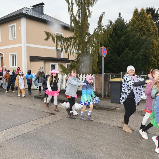 Eine Gruppe von Kindern in Kostümen geht vor einem Haus mit Schornstein und weißer Hecke die Straße entlang.