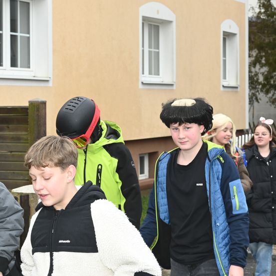 Eine Gruppe von Kindern geht vor einem Gebäude. Ein Junge trägt einen schwarzen Helm. Ein anderer trägt eine Perücke. Sie sind alle warm für das Wetter gekleidet.