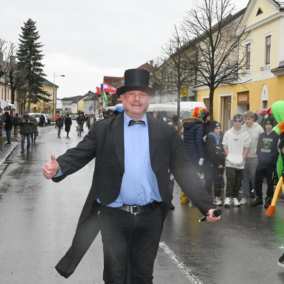 Ein Mann in einem Zylinder und einem blauen Hemd lächelt und gibt an einer nassen Straße einen Daumen hoch, mit Gebäuden und Menschen im Hintergrund.