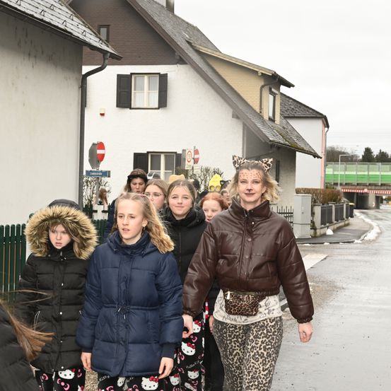 Eine Gruppe von Kindern und Erwachsenen in Winterkleidung geht auf einer verschneiten Straße. Eine Frau mit einem Leopardenprint-Rock und einer Gesichtsmaske führt sie an.