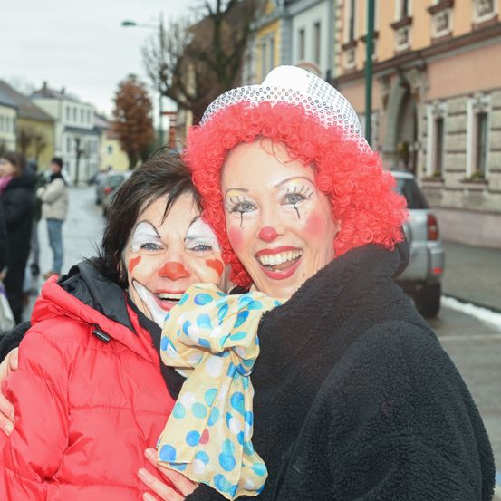 Zwei Frauen, die als Clowns verkleidet sind, umarmen sich auf einer Stadtstraße. Eine Frau hält ein buntes Tuch und beide tragen Make-up und Perücken.