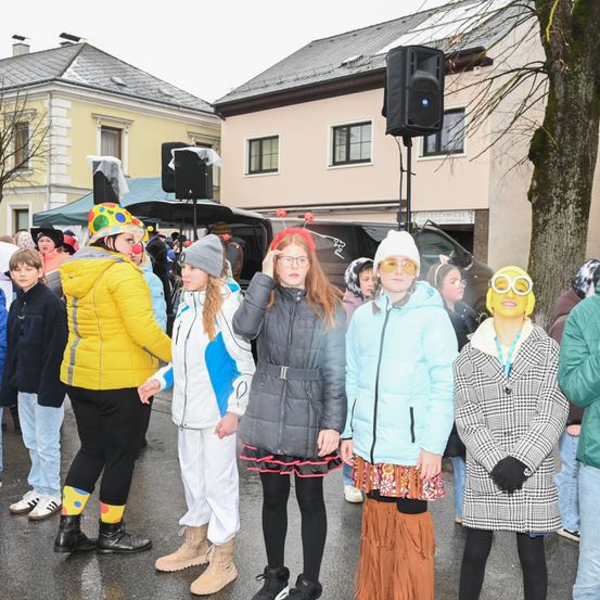 Eine Gruppe von Menschen in bunten Kostümen steht in einer Reihe auf einer Straße. Das Wetter ist kalt, und sie tragen Winterkleidung. Im Hintergrund befindet sich ein Baum und ein Gebäude mit Lautsprechern.