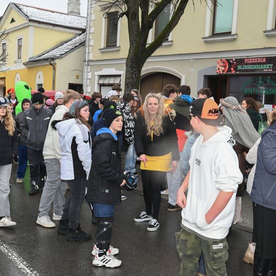 Eine Gruppe von Menschen in Winterkleidung, einige mit Gesichtsbemalung, stehen auf einer nassen Straße vor einem Gebäude mit einem Schild, das Pizzeria Blindenmarkt liest.