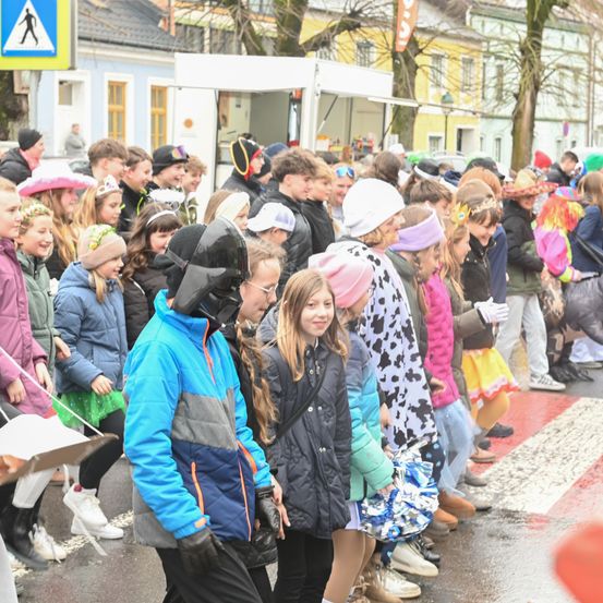 Eine Menschenmenge, überwiegend Kinder, steht auf einer nassen Straße. Sie tragen verschiedene Mützen und Kostüme. Einige halten Papiere, und eine Person vorne trägt eine blaue Jacke mit einer Maske. Bäume und Gebäude sind im Hintergrund.