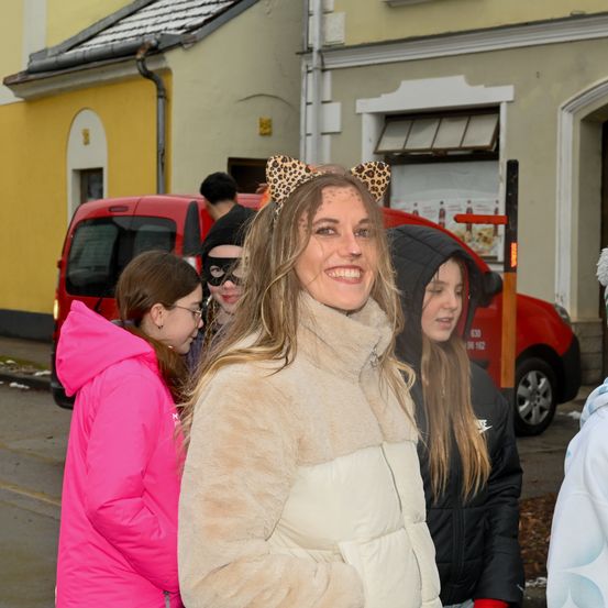 Eine Gruppe von Menschen steht draußen. Eine Frau trägt Leoparden-Katzenohren und lächelt. Hinter ihr trägt eine andere Frau eine Maske. Ein roter Van ist geparkt.