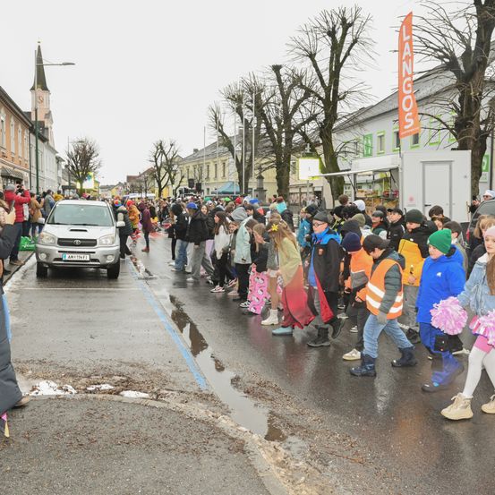 Eine Menschenmenge in Winterkleidung steht auf einer nassen Straße, mit einem silbernen Toyota-Auto auf der Seite geparkt und einem Gebäude mit grünem Vordach im Hintergrund.