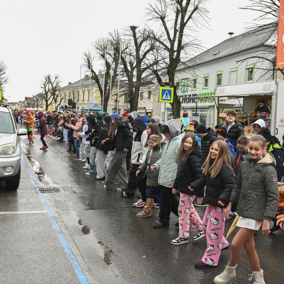 Eine Gruppe von Menschen in bunten Kostümen und Winterkleidung steht in einer Schlange auf einer nassen Straße, wahrscheinlich bei einem Umzug. Gebäude säumen die Straße, mit einem Imbisswagen auf der Seite.
