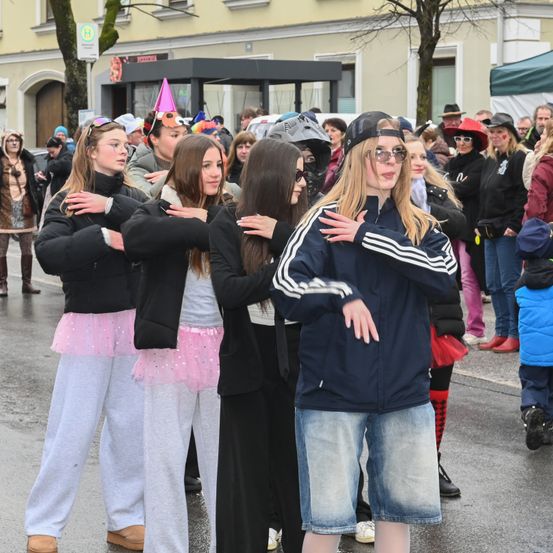 Eine Gruppe junger Leute steht auf einer Straße, einige in Kostümen, darunter rosa Tüllröcke und Brillen. Sie stehen in einer Reihe und blicken in die Kamera, mit einer vielfältigen Menge hinter ihnen. Der Hintergrund zeigt ein Gebäude mit einer Bushaltestelle und einem Baum.