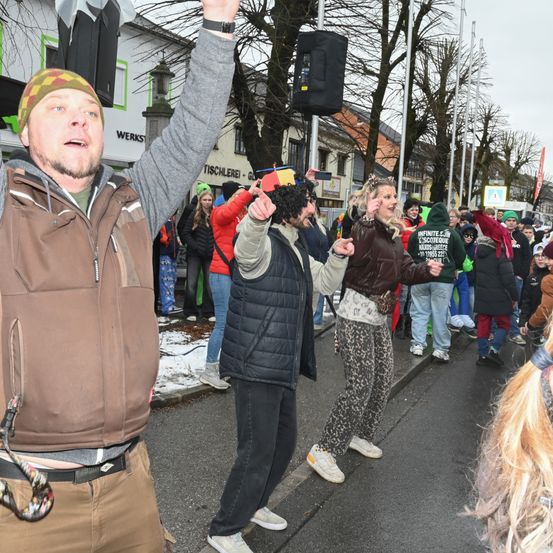 Eine Straßenszene mit Schnee auf dem Boden. Menschen in verschiedenen Kostümen und Kleidungen gehen und tanzen. Ein Mann in einem braunen Mantel hebt seine Hände, während ein anderer in einem Leoparden-Print-Outfit zusieht.