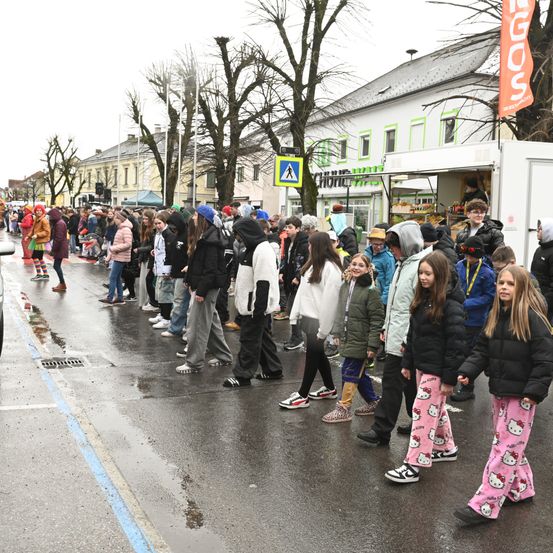 Eine Gruppe von Menschen, darunter Kinder, steht in einer Reihe auf einer nassen Straße, möglicherweise in Erwartung einer Veranstaltung. Es gibt Bäume, Gebäude und einen Imbisswagen in der Nähe.