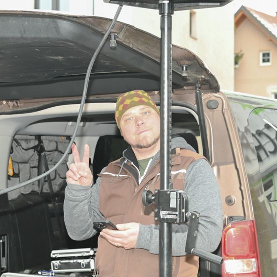 Ein Mann mit Hut und Jacke steht am offenen Kofferraum eines Fahrzeugs, hält ein Handy und macht ein Friedenszeichen.