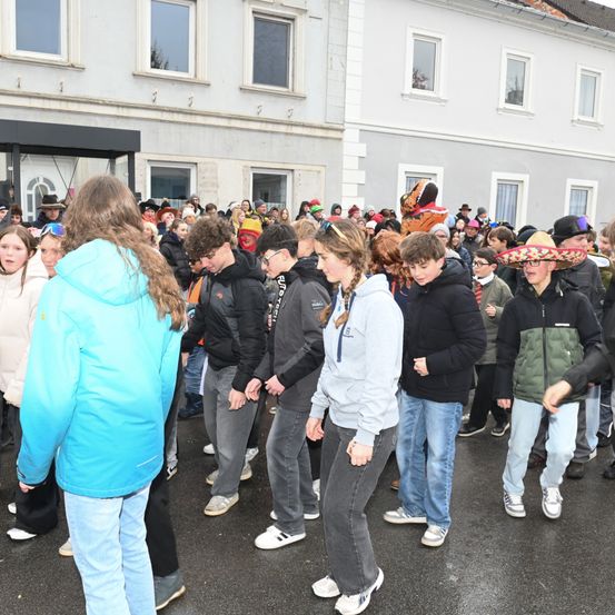 Eine Menschenmenge, teilweise in Kostümen, versammelt sich auf einem Außengelände, vermutlich für eine Veranstaltung. Eine Person trägt ein Dinosaurierkostüm. Sie stehen in einer Reihe, möglicherweise auf etwas wartend. Vor ihnen steht ein Gebäude mit mehreren Fenstern. Einige Menschen tragen Hüte, Brillen und Turnschuhe.