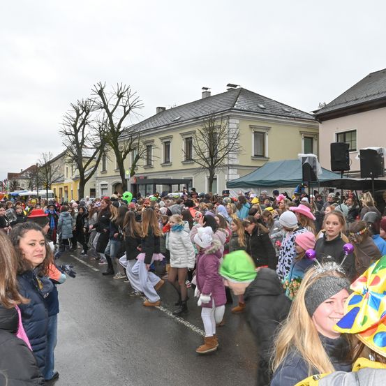 Eine Menschenmenge in festlichen Hüten und Kostümen versammelt sich auf der Straße, wahrscheinlich für eine Parade. Die Gebäude im Hintergrund haben viele Fenster, und es gibt Bäume davor. Lautsprecher und Zelte sind aufgestellt, und der Himmel ist bewölkt.