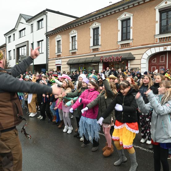 Eine Gruppe von Menschen, darunter Kinder in Kostümen, tanzt auf der Straße vor einem Gebäude mit einem Schild, das 'Fischer' steht.