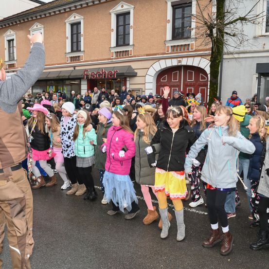 Eine große Gruppe von Kindern und Erwachsenen versammelt sich vor einem Gebäude, wahrscheinlich für ein Festival. Die Kinder tragen farbenfrohe Kostüme und Stiefel, und einige tragen Hüte. Der Erwachsene auf der linken Seite hebt seine Hand. Das Gebäude hat ein Schild mit der Aufschrift Fischer.