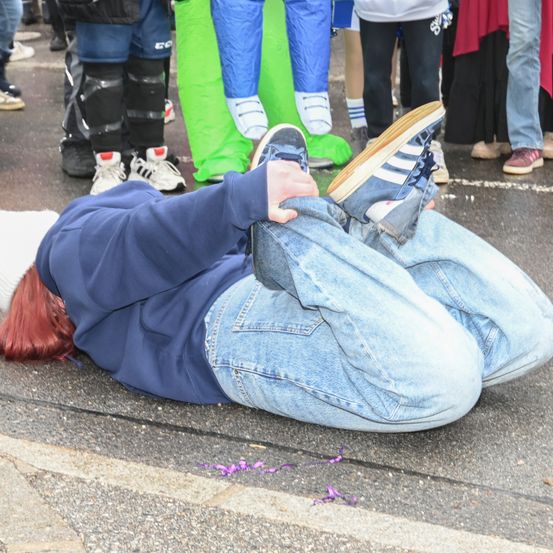 Eine Person in blauer Jeans und einem Sweatshirt liegt mit hochgereckten Schuhen auf dem Boden. Dahinter stehen verschiedene Menschen in verschiedenfarbiger Kleidung.