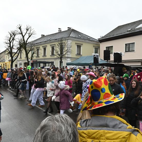 Eine große Menschenmenge, viele in Kostümen gekleidet, versammelt sich auf einer Straße, vermutlich für eine Parade. Gebäude säumen die Straße, und in der Mitte ist ein Zelt aufgestellt. Einige Leute gehen, während andere stehen und zusehen.