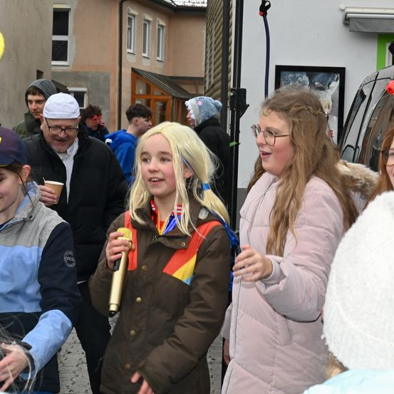 Eine Gruppe von Menschen, darunter zwei Mädchen und Erwachsene, versammelt sich im Freien. Ein Mädchen hält ein Mikrofon und lächelt, während das andere Mädchen vorausblickt. Ein Mann mit Brille steht hinter ihnen. Gebäude mit Fenstern und ein geparktes Auto sind im Hintergrund zu sehen.