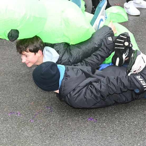 Zwei Jungen liegen auf dem Boden, einer mit erhobenen Füßen, während ein anderer seinen Kopf auf die Schulter seines Freundes legt. Ein grünes aufblasbares Objekt ist in der Nähe.