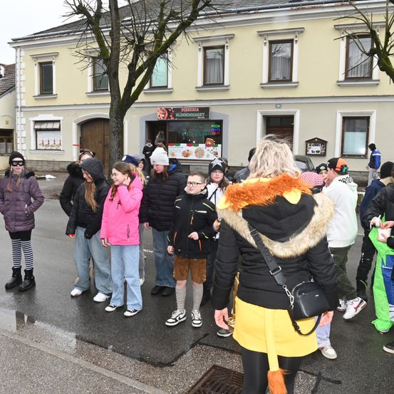Eine Gruppe von Menschen in Winterkleidung versammelt sich auf einer nassen Straße vor einem Gebäude mit einem Schild, auf dem Pizza Blinoma steht. Eine Frau in einem gelben Rock und Fuchsohr-Kopfband steht in der vordersten Reihe.
