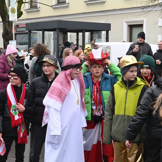 Eine Gruppe von Menschen in verschiedenen Kostümen, darunter ein weißes Gewand und ein rot-weißes Halstuch, steht auf einer Straße. Einige tragen Brillen und Mützen. Im Hintergrund befinden sich Gebäude und ein weißer Lieferwagen.