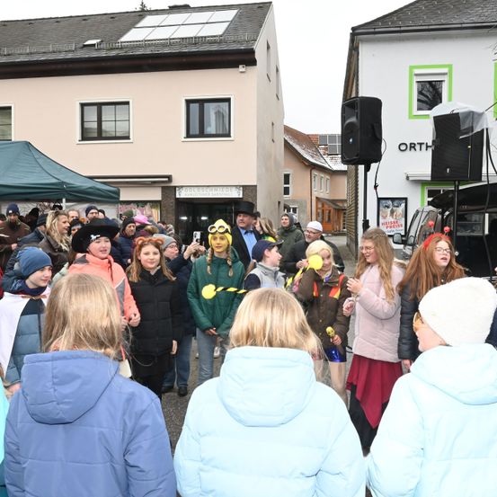 Eine Menschenmenge, darunter Kinder in Kostümen, versammelt sich vor einem Gebäude mit Solarpanels auf dem Dach. Eine Person mit gelber Maske und Schutzbrille steht in der Mitte.