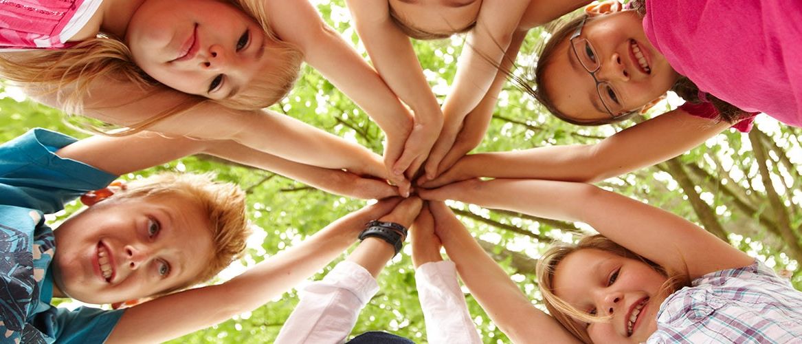 Eine Gruppe lächelnder Kinder hält unter einem Baum Händchen. Sie sehen fröhlich aus und tragen Freizeitkleidung.