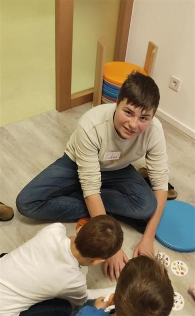 Ein junger Mann sitzt mit zwei Kindern auf dem Boden. Er lächelt und spielt mit ihnen. Es gibt Teller und Spielzeug auf dem Boden.