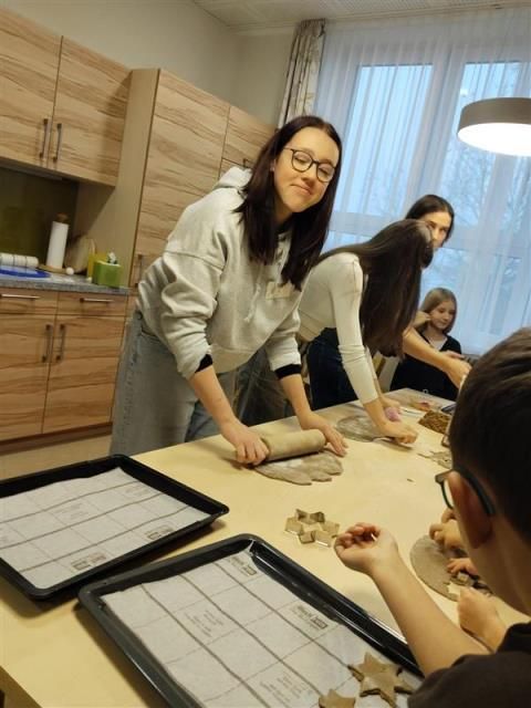 Mehrere Frauen und Kinder sind in einer Küche und rollen Teig auf einem Tisch aus. Tabletts mit Backpapier stehen auf dem Tisch, und eine Frau rollt Teig auf einem Nudelholz.