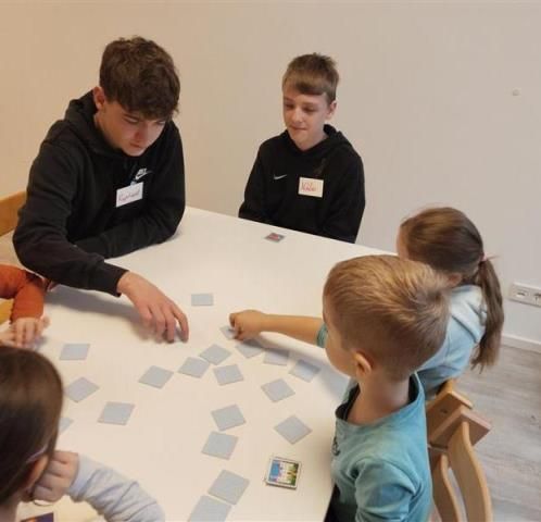 Eine Gruppe von Kindern und zwei jungen Männern sitzen um einen Tisch. Der Tisch ist mit Karten bedeckt und die Kinder spielen ein Spiel.