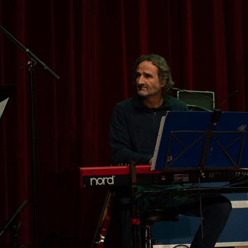 Ein Mann sitzt auf einem Schemel und spielt Klavier mit einem roten Nord-Keyboard vor ihm. Er blickt auf einen blauen Notenständer.