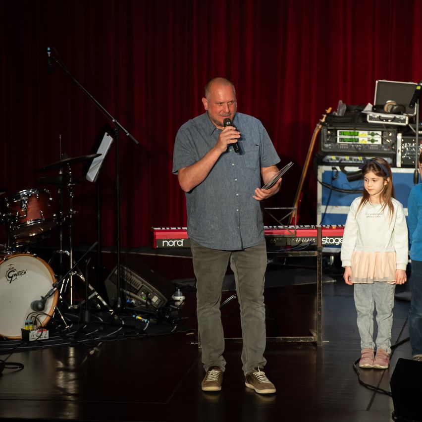 Ein Mann steht auf der Bühne, hält ein Mikrofon und schaut eine neben ihm stehende Mädchen an. Dahinter befinden sich ein Schlagzeug, ein Lautsprecher und ein Keyboard.